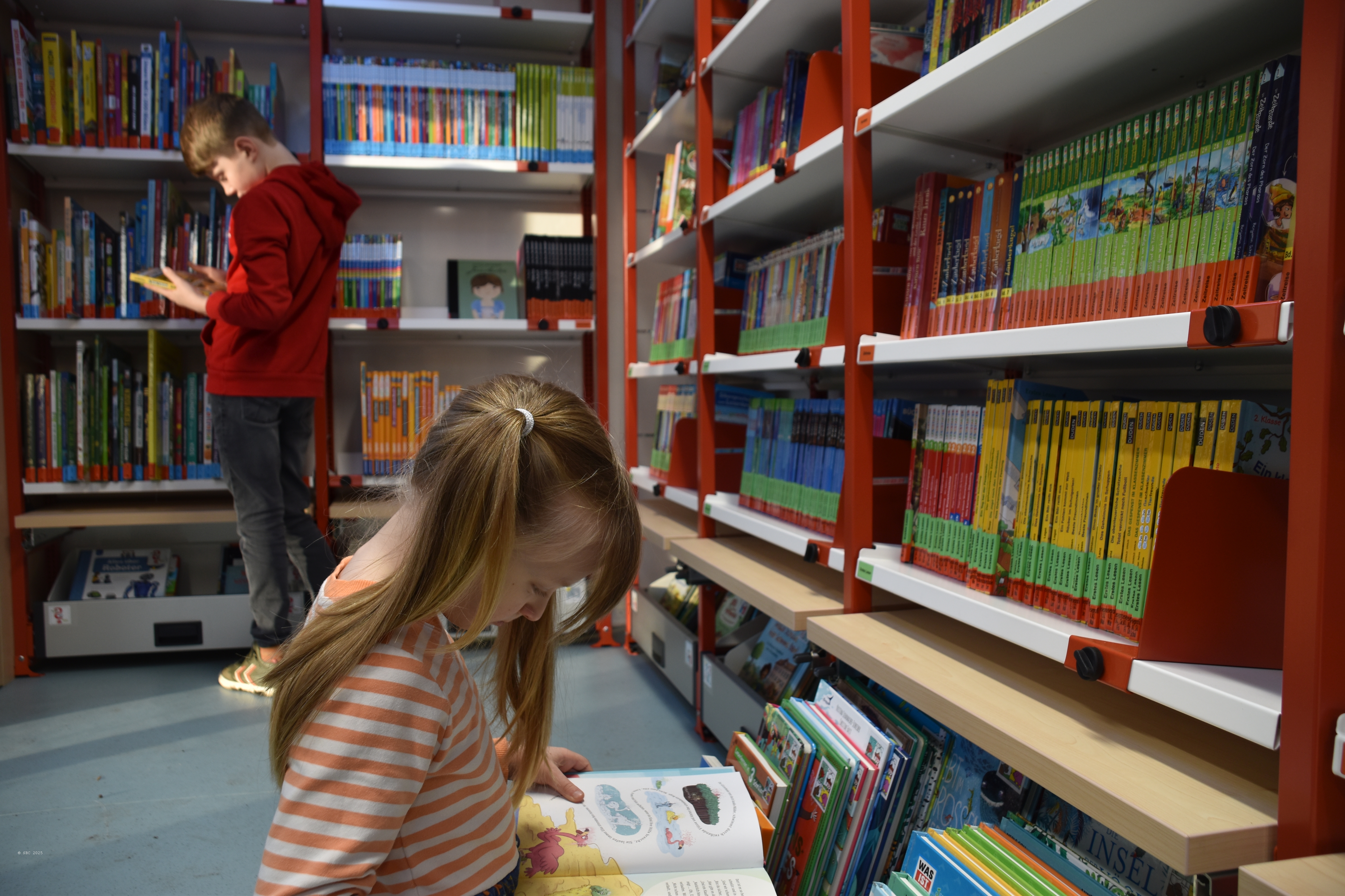 Foto zeigt das Innenraum mit vielen bunten Bücherregalen und einem Jungen und ein Mädchen, die je ein Buch anschauen und dabei vertieft sind