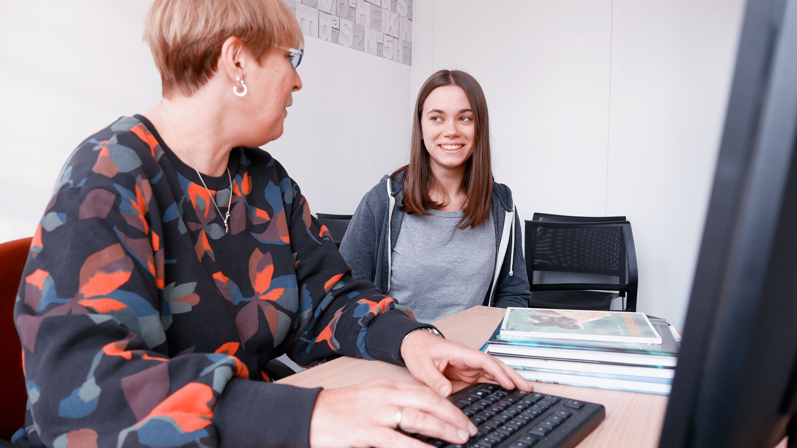 Frau am PC unterhält sich mit Schülerin im Büro