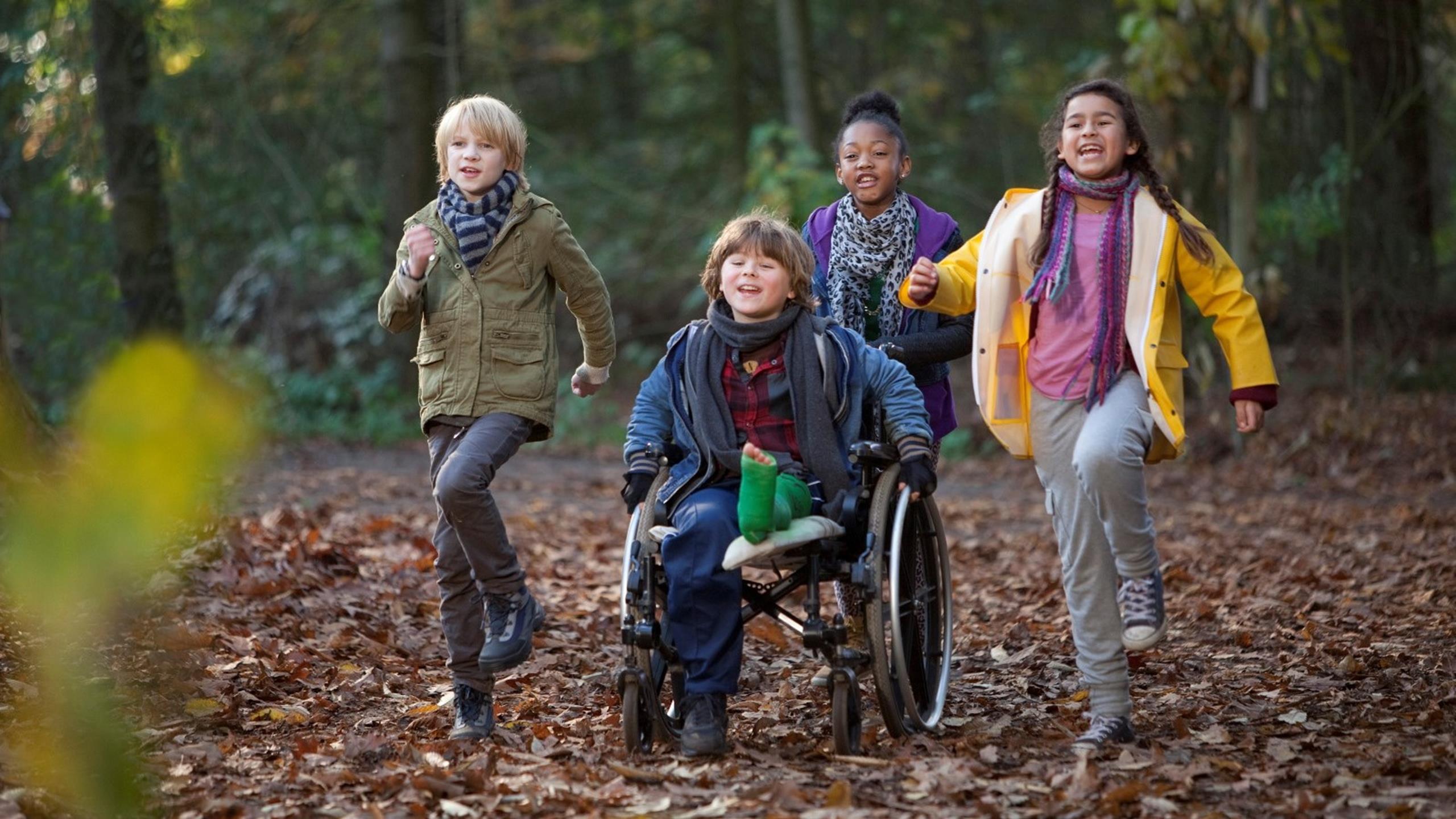 Vier Kinder rennen im Wald auf den Betrachtenden zu, ein Junge sitzt im Rollstuhl, er hat ein Bein eingegipst und wird von einem Mädchen geschoben